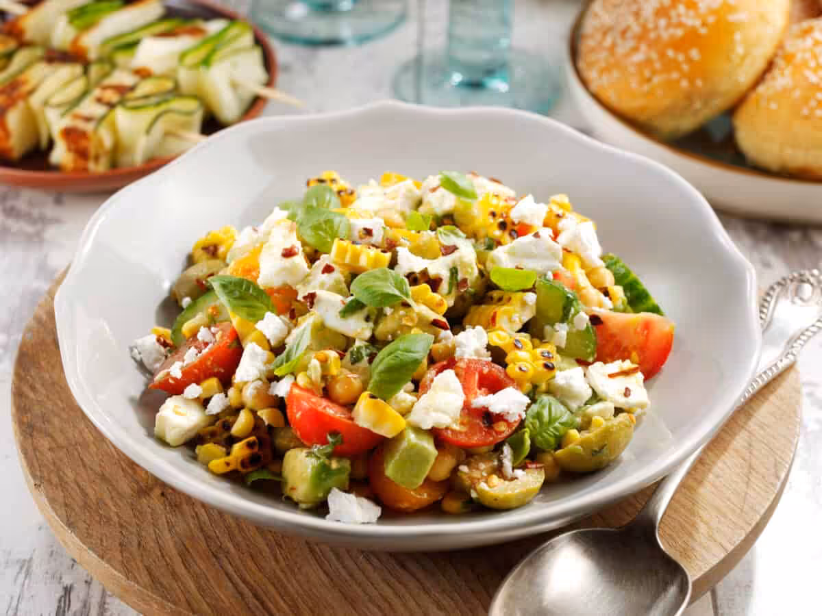 A white bowl filled with a feta salad recipe including charred corn, avocado and green leaves, on a wooden table