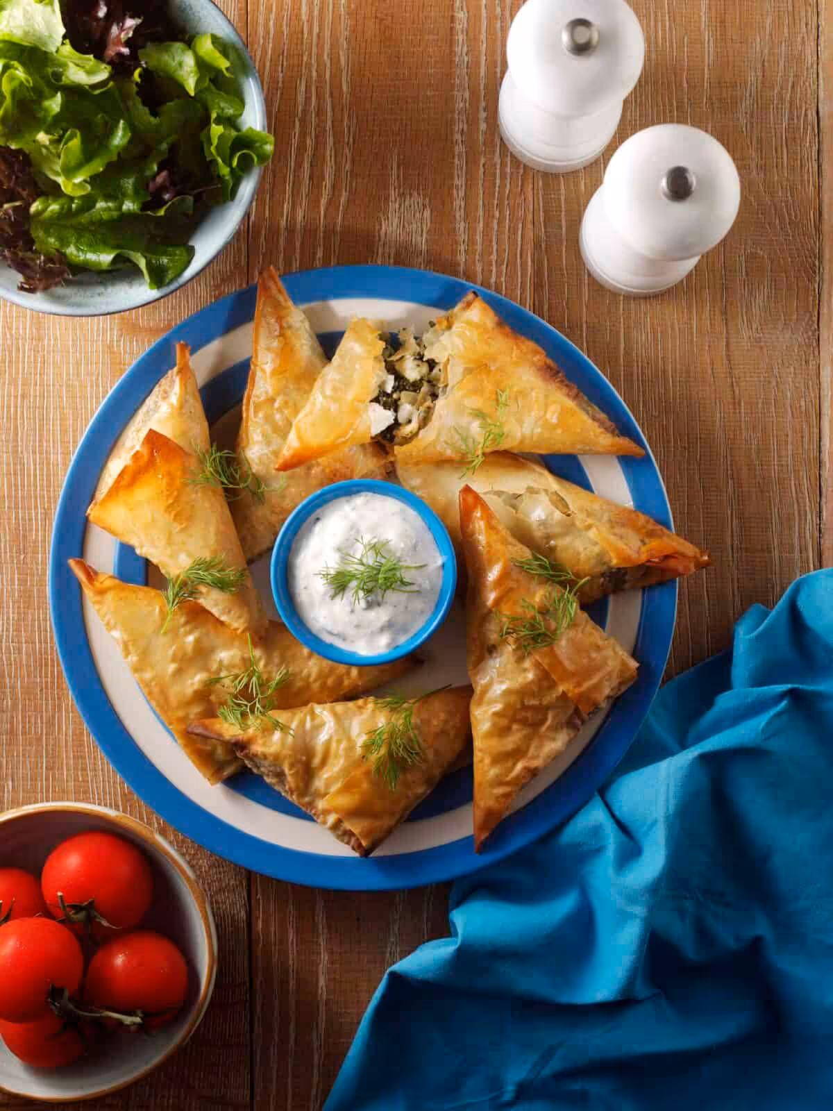 Spinach and feta parcels on a blue and white place surrounded by salad, a blue napkin and a pack of feta cheese