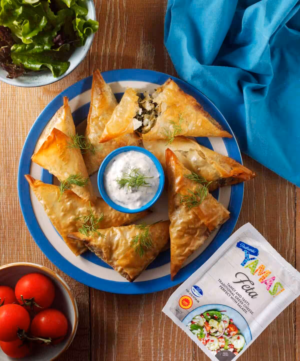 Spinach and feta parcels on a blue and white place surrounded by salad, a blue napkin and a pack of feta cheese with a packet of YAMAS! Feta cheese