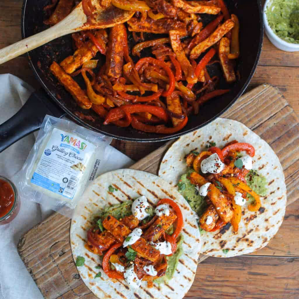 A frying pan full of vegetables fried in fajita mix, with tortillas lying flat in front of them with a filling of vegetables and halloumi.