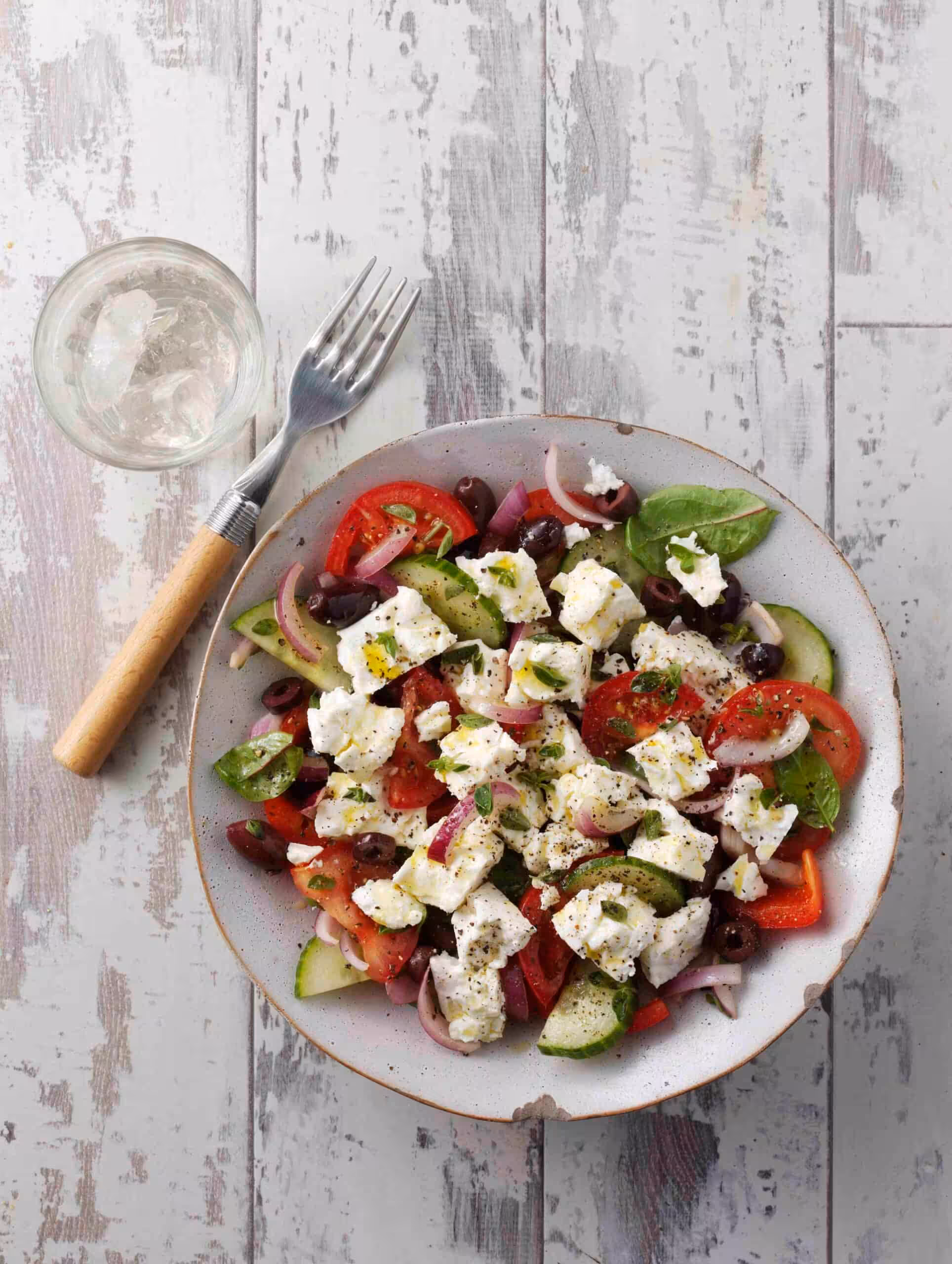 A bowl of greek salad (tomatoes, cucumbers, olives and feta) on a white boarded table