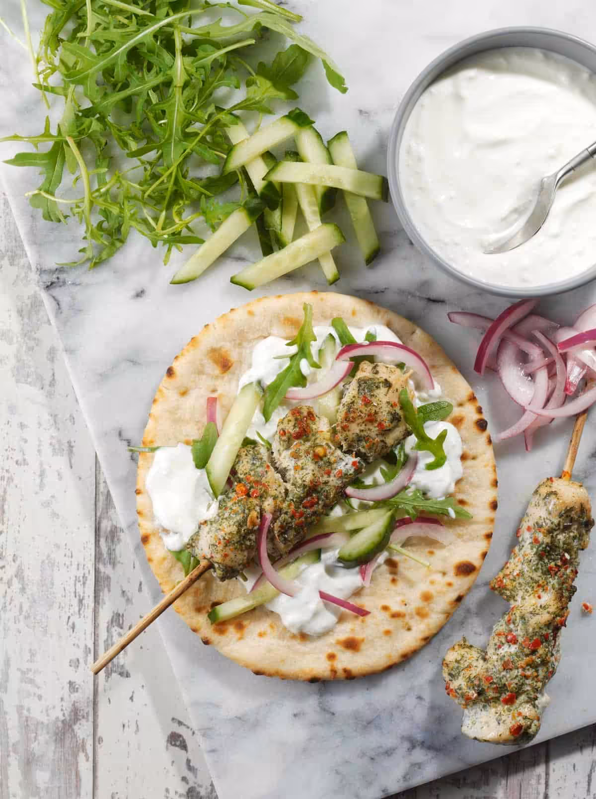 A flatbread on a chopping board with whipped feta, salad, and chicken skewers on top, with ingredients surrounding it.