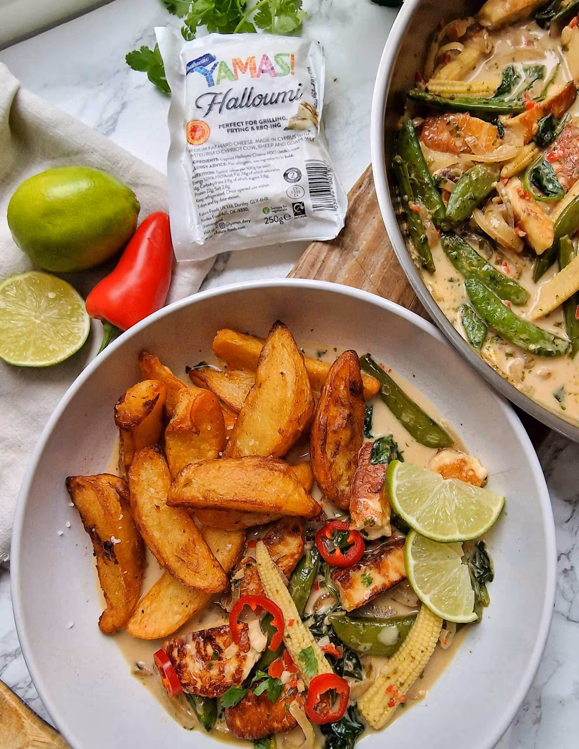 A white bowl containing Thai green curry, chips and lime, surrounded by a pan of curry, limes, chillis and a pack of halloumi