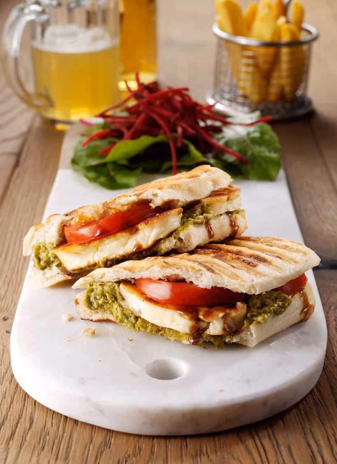 A toasted panini filled with a layer of pesto adn slices of halloumi and tomatoes. On a white marble serving board with a side of salad.
