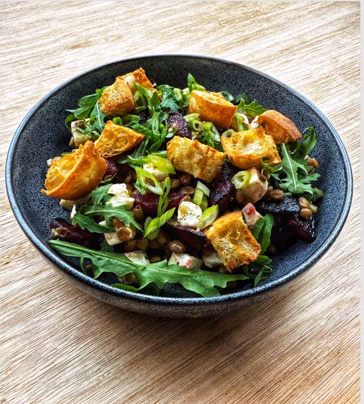 A dark bowl on a light wooded table, filled with a green rocket salad, topped with dark red beetroot and feta cubes.