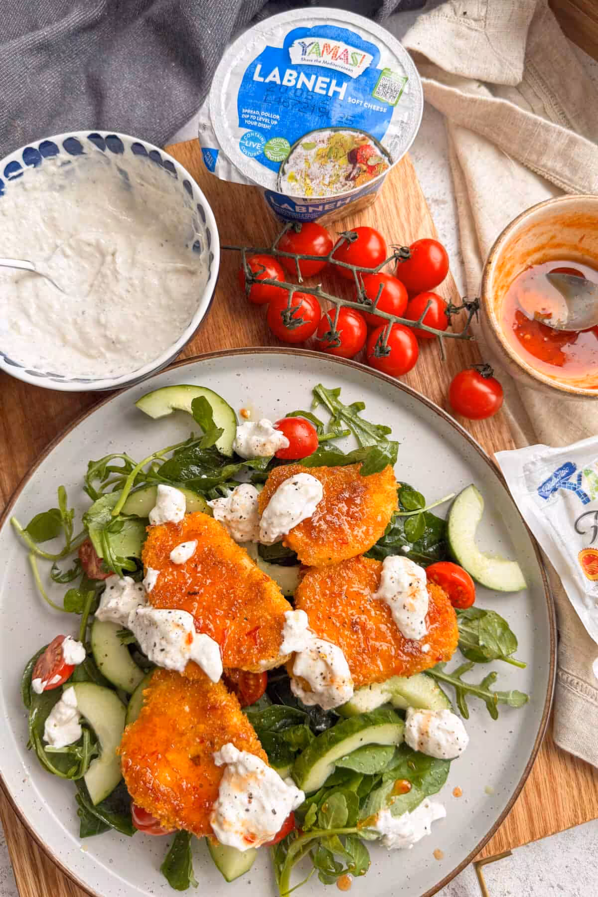 Stone Marble table with a speckled light grey bowl filled with a salad topped with triangle breaded YAMAS! Halloumi with YAMAS! Labneh dolloped over. This is on a wooden chopping board with loose red tomatoes on the side with a bowl of YAMAS! Labneh and a pot of the branded pot, with a smaller rustic pot with hot honey with a spoon going out of frame.. There is a rustic light cream kitchen towel underneath holding the hot honey pot.