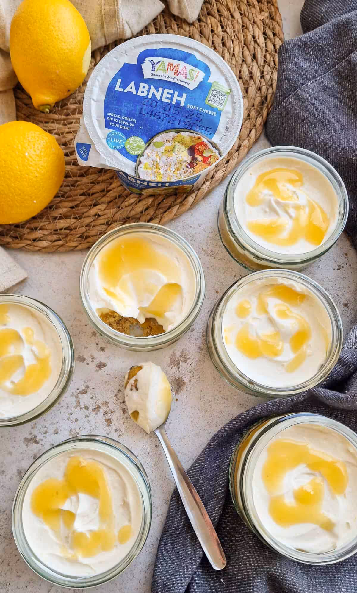 A light grey marble effect table shot from above with 6 small glass pots filled with cheesecake with lemon drizzled on top. One pot in the middle has a chunk taken out with a spoon resting next to it with crumbs of the cheesecake base. Next to the pots on a straw mat with 2 whole lemons and a pack of YAMAS! Labneh on the side of the shot.